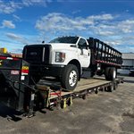 Ford Medium-Duty Stake Truck — A white Ford medium-duty truck with tall black stake sides is secured on a step-deck.