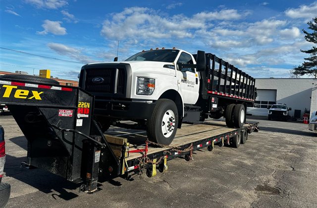 Ford Medium-Duty Stake Truck — A white Ford medium-duty truck with tall black stake sides is secured on a step-deck.
