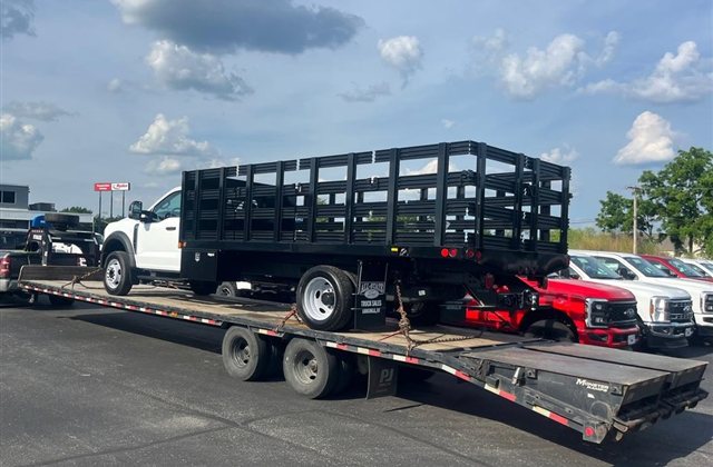 Ford Super Duty Stake-Body Truck — A white Ford Super Duty stake-body truck is photographed from two sides on a step-deck trailer. View 1.