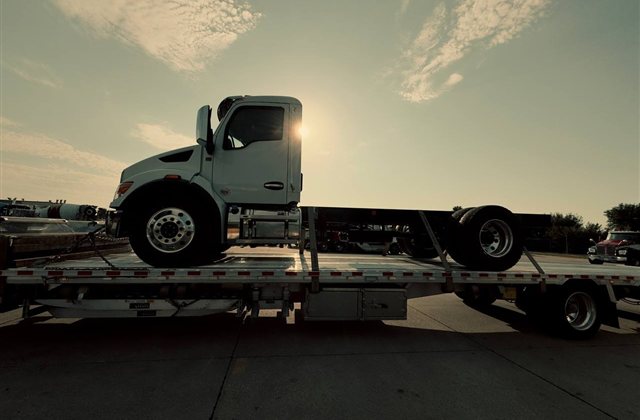 White Truck Chassis — A white conventional truck cab and exposed chassis are photographed on a transporter in low light. View 1.